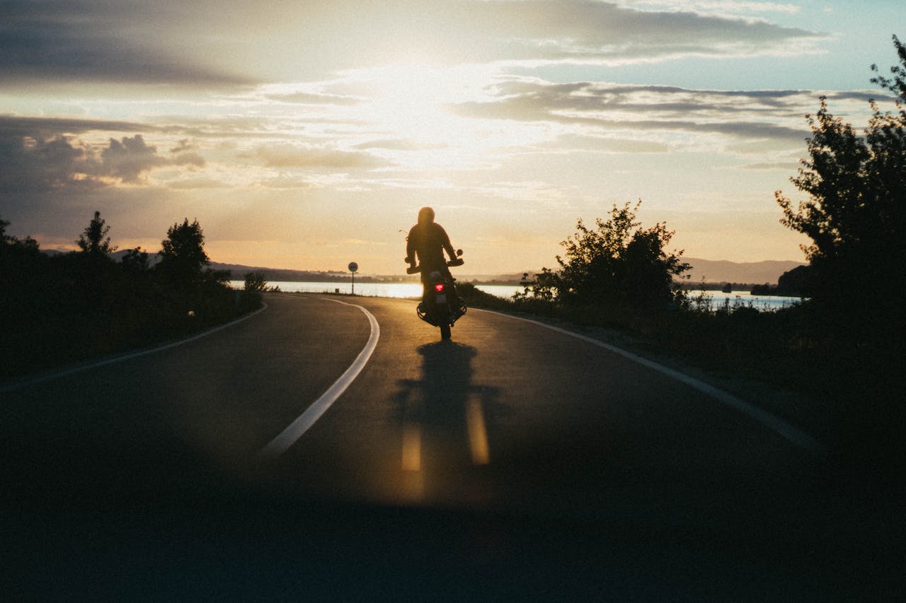 heros-img Silhouette of a motorcyclist riding on a scenic road during a beautiful sunset, emphasizing freedom and adventure.